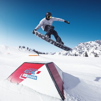 Snowboarder springt mit Grab über ein Obstacle im RIDE Ski amadé Snowpark, blauer Himmel und Berge im Hintergrund.