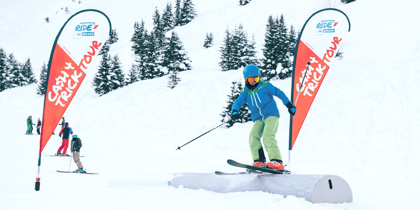 Young child in blue jacket and green pants skis on a box during Cash4Tricks event, framed by two red promotional flags.