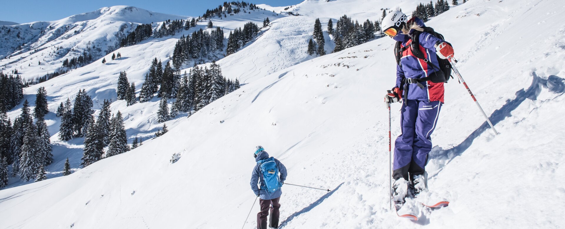 Zwei Freerider stehen leicht versetzt und mit dem Rücken zueinander auf einem Hang und sehen ins Tal hinunter