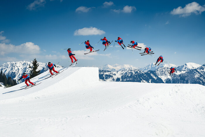 Freeski trick sequence over kicker in snowpark Sequence shows freeskier in red-blue outfit jumping over kicker in snowpark with snowy alpine peaks in background.