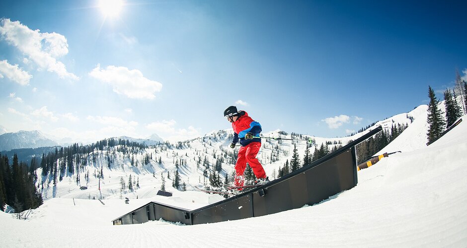 Freeskier in rot-blauem Outfit slidet auf Rail im Snowpark, Sonnenlicht und verschneite Berge bilden die Kulisse.