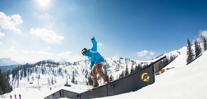 Snowboarder in blauem Hoodie slidet bei Sonne über Rail im Snowpark, verschneite Berge und Wald im Hintergrund.