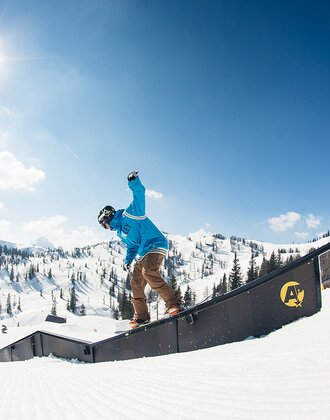 Snowboarder in blauem Hoodie slidet bei Sonne über Rail im Snowpark, verschneite Berge und Wald im Hintergrund.