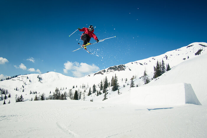 Freeskier skis mid-air above kicker in snowpark Freeskier in red jacket executes trick above kicker with blue sky and snowy peaks in background of snowpark.