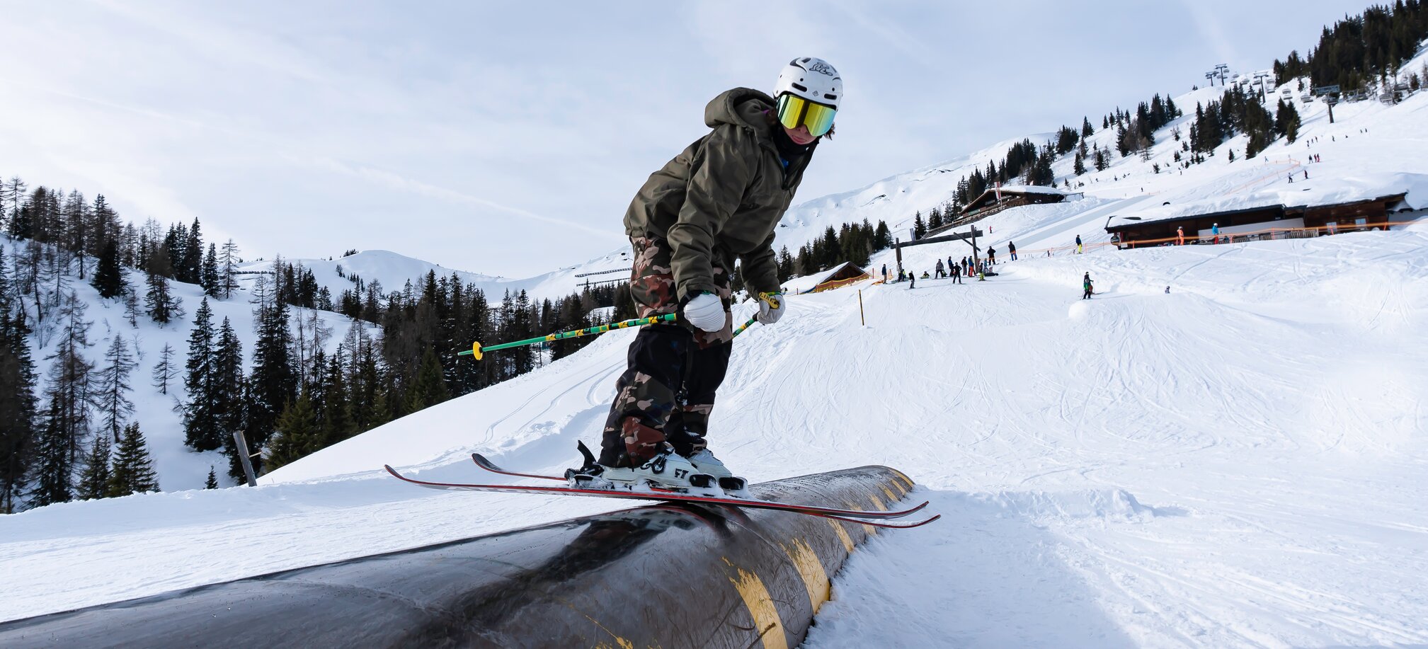 Matej Svancer slidet auf schwarz-gelbem Tube im Freeski-Park, mit Camouflage-Hose und Helm, Zuschauer im Hintergrund.