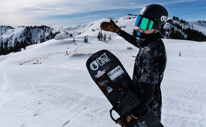 Snowboarder zeigt auf den Snowpark Kings Park Hochkönig Snowboarder mit Helm und Brille zeigt lachend auf den Kings Park Hochkönig mit Obstacles im verschneiten Gelände.