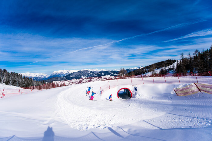 Kids ski through snow tunnel on funslope under bright blue sky Three kids ski through a snow tunnel on the funslope, with sunshine and a mountain panorama in the background.