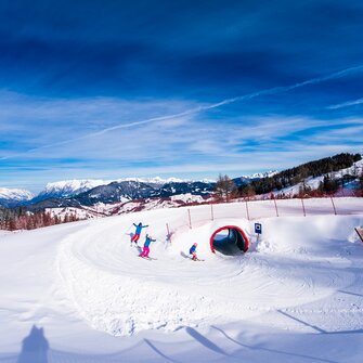 Three kids ski through a snow tunnel on the funslope, with sunshine and a mountain panorama in the background.