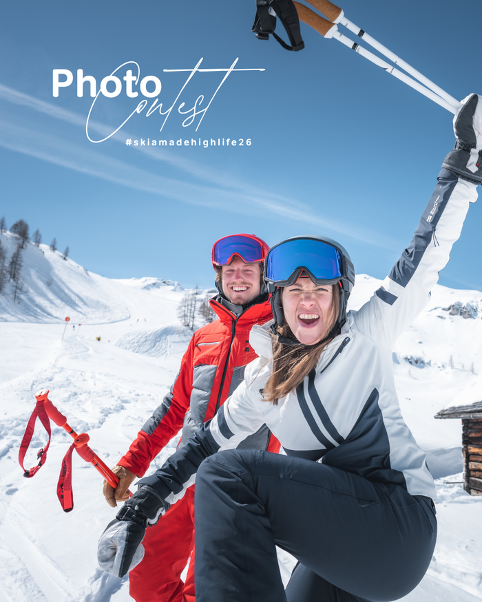 Zwei Skifahrer in roter und weisser Skikleidung posieren mit Stöcken im Schnee vor Bergen und blauem Himmel