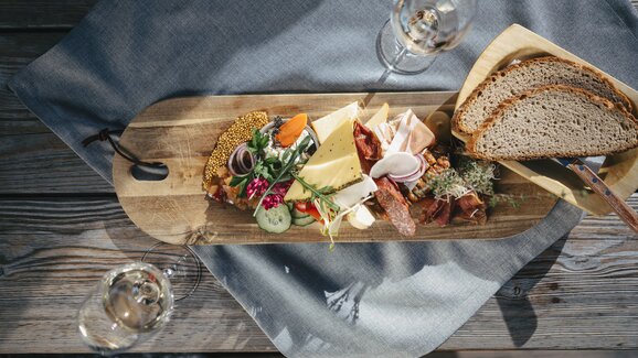 Cheese, cold cuts, bread, vegetables and herbs arranged on wooden board with two glasses of white wine | © Ski amadé
