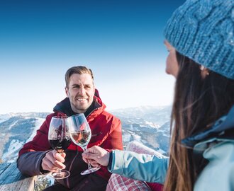 A man in a red ski jacket smiles and clinks glasses with a woman in a blue cap and blue ski jacket as they sit at a wooden table