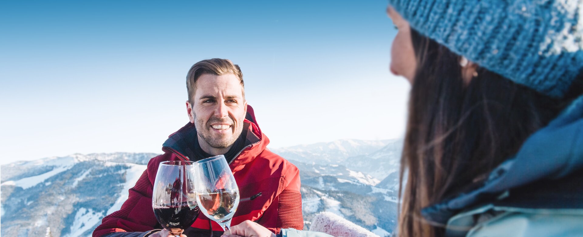 A man in a red ski jacket smiles and clinks glasses with a woman in a blue cap and blue ski jacket as they sit at a wooden table