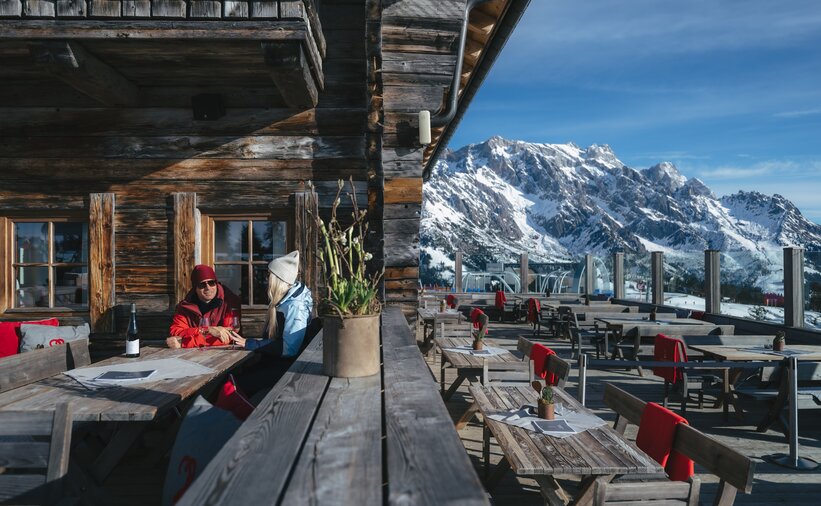 Empty sunny terrace with wooden tables and snowy mountains, two people sitting by the hut | © Ski amadé