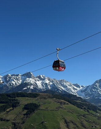 Rote Gondel der Abergbahn schwebt im Sommer vor grüner Landschaft und schneebedecktem Hochkönig-Massiv bei blauem Himmel. | © Aberg - Hinterthal - Bergbahnen AG