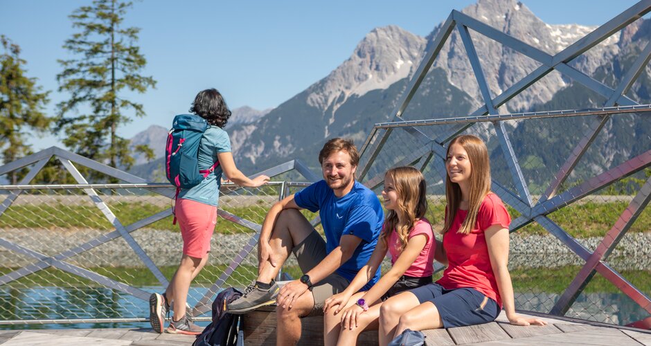 Vier Wanderer rasten an einer Plattform am Prinzensee in Maria Alm mit Panoramablick auf die Berge des Hochkönigs. | © Roland Haschka