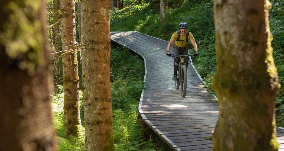 Ein Mountainbiker mit Helm und Protektoren fährt auf einem kurvigen Holzsteg durch einen dichten, sonnendurchfluteten Wald. | © Roland Haschka