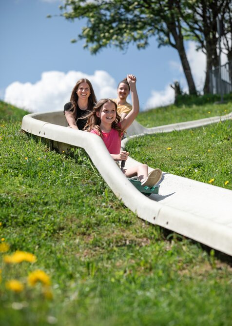 Lachendes Mädchen fährt mit Familie auf Sommerrodelbahn den grünen Hang hinunter, Sommerwiese im Vordergrund. | © Hochkönig / Roland Haschka