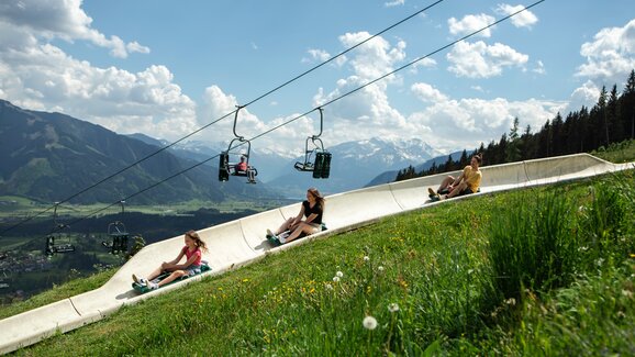 Drei Personen rodeln auf der Sommerrodelbahn Biberg durch Wiesenlandschaft, darüber Sessellift und Bergpanorama im Hintergrund. | © Hochkönig / Roland Haschka