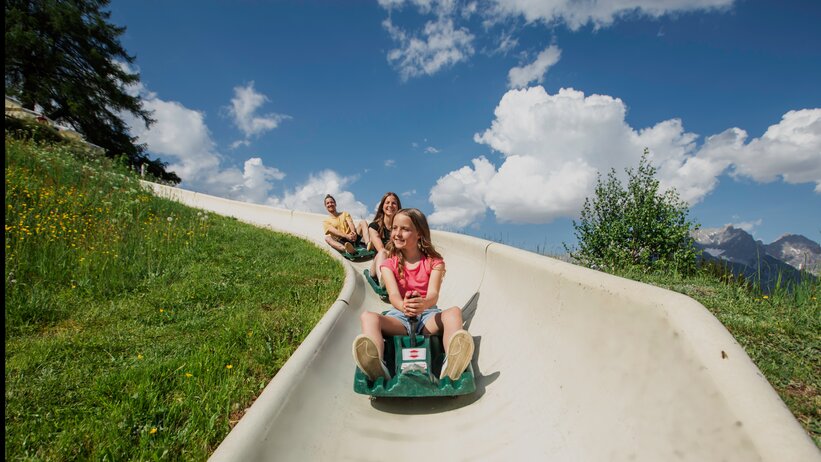 Mädchen rodelt mit zwei Erwachsenen hinter sich die Sommerrodelbahn Biberg hinunter, grüne Wiese, blauer Himmel. | © Hochkönig / Roland Haschka