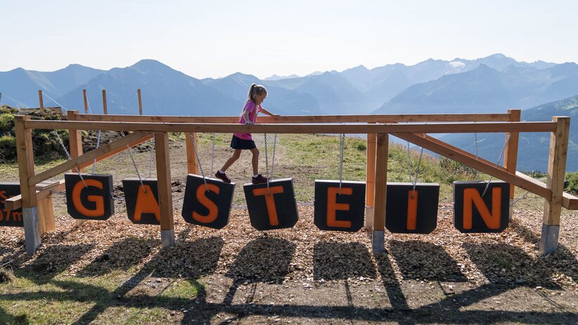 Mädchen balanciert über Spielgerät mit GASTEIN-Buchstaben vor Bergpanorama im Gasteinertal | © Gasteiner Bergbahnen AG