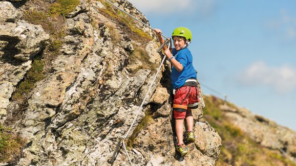 Junge mit Helm und Gurt klettert an gesichertem Felsen bei Sonnenschein in alpiner Landschaft | © Gasteiner Bergbahnen AG