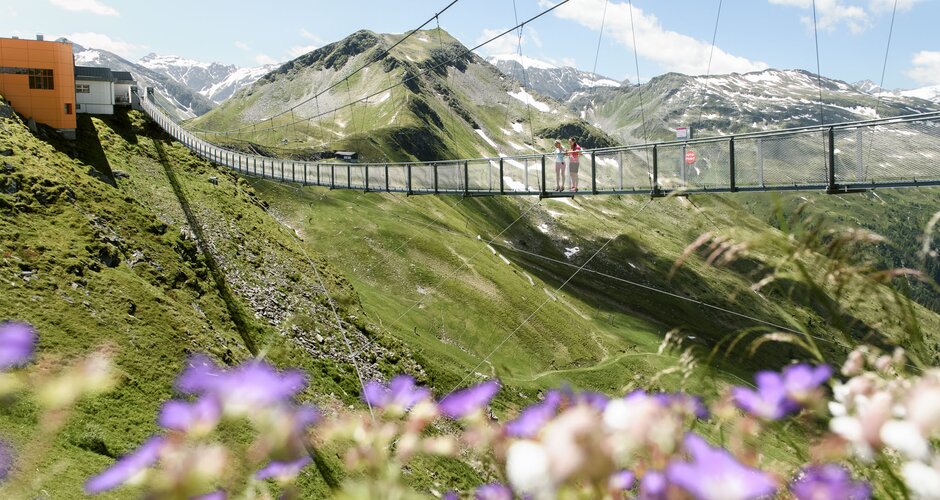 Zwei Personen stehen auf der Hängebrücke am Stubnerkogel, im Vordergrund blühen violette Sommerblumen | © Gasteiner Bergbahnen, Marktl Photography