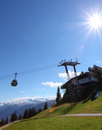 Gondel fährt bei Sonnenschein zur Bergstation, grüne Almwiese und verschneite Bergkulisse im Hintergrund, blauer Himmel. | © Großarler Bergbahnen