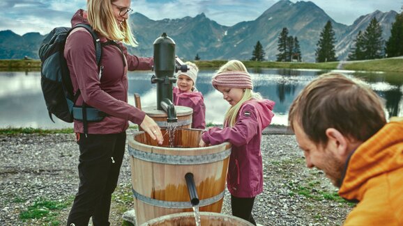 Family moment at the water pump in Großarl | © Großarler Bergbahnen Family with kids pumping water from wooden barrels by a lake in Großarl, mountains and clouds reflecting in the background. | © Großarler Bergbahnen