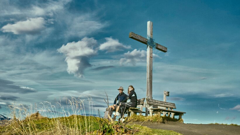 Resting at summit cross in Großarl | © Großarler Bergbahnen Two people rest on a bench by the summit cross in Großarl, enjoying the view beneath a dramatic, partly cloudy sky. | © Großarler Bergbahnen