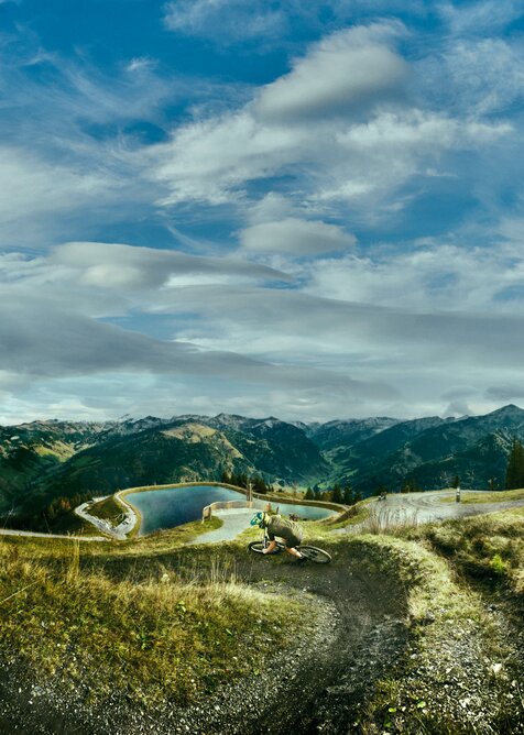 Mountain biking with panoramic view in Großarl | © Großarler Bergbahnen Mountain biker on winding trail overlooking reservoir, forested slopes and a sky full of dramatic clouds. | © Großarler Bergbahnen