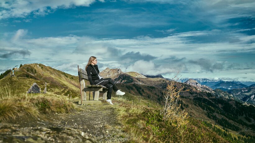 Frau sitzt auf einer Bank am Bergweg in Großarl und blickt über eine herbstliche Hügellandschaft mit dramatischer Wolkenkulisse. | © Großarler Bergbahnen