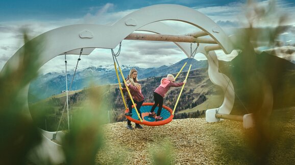 Zwei Kinder spielen auf einer großen Nestschaukel vor Bergkulisse in Großarl, Holzhackschnitzel am Boden, Wolken am Himmel | © Großarler Bergbahnen