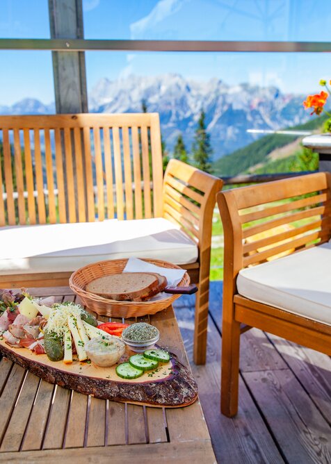 Holztisch mit Brettljause, Brotkorb und Menükarte auf sonniger Terrasse mit Blick auf Berge und blühende Geranien. | © René Eduard Perhab