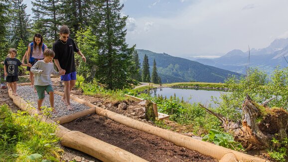 Familie mit Kindern barfuß auf einem Naturpfad mit Kies und Erde am Hauser Kaibling, im Hintergrund ein Bergsee und Alpenpanorama. | © René Eduard Perhab