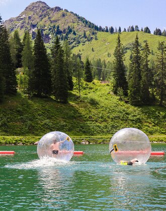 Zwei Kinder in transparenten Zorbing-Bällen auf einem See vor grüner Bergkulisse. | © Hauser Kaibling