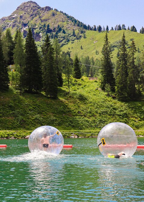 Zwei Kinder in transparenten Zorbing-Bällen auf einem See vor grüner Bergkulisse. | © Hauser Kaibling