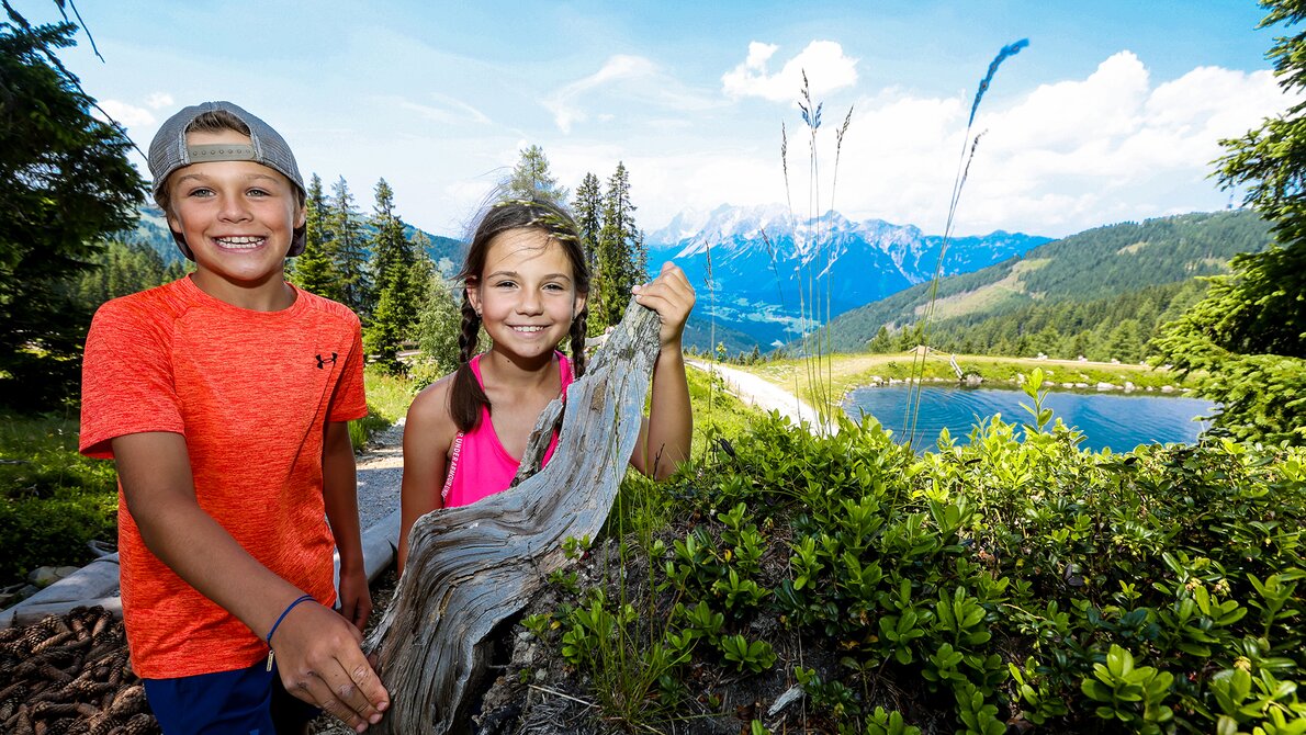 Zwei Kinder lächeln vor einem Baumstamm am Schafsinn-Teich mit Bergblick im Sommer. | © Stefan Pajman