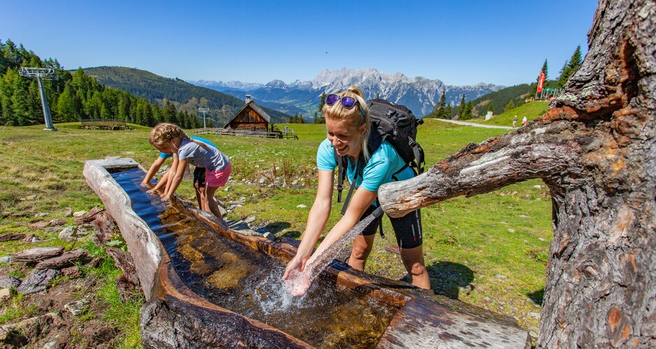 Frau und Mädchen erfrischen sich an einem Wasserbrunnen aus Holz beim Wandern mit Hüttenblick in alpiner Berglandschaft. | © Hauser Kaibling
