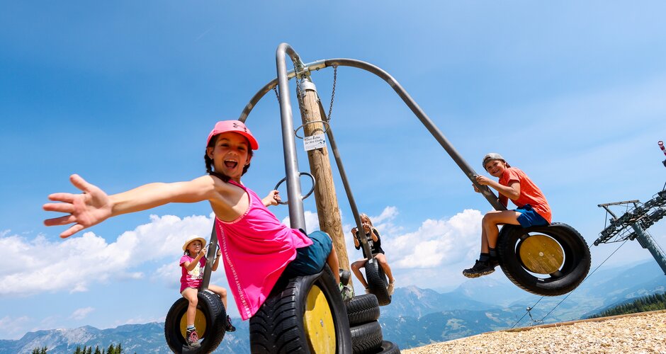 Kinder schwingen auf einem Karussell aus Reifen vor Bergkulisse, lachen und strecken die Arme fröhlich in die Kamera. | © Stefan Pajman
