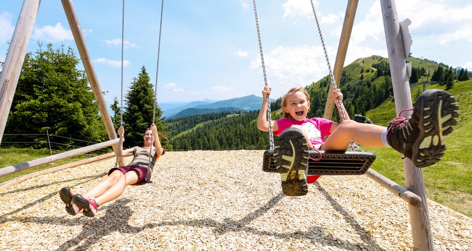 Kind und Frau schaukeln lachend auf Holzschaukeln mit Blick auf grüne Berge, Waldlandschaft und sonnigen Himmel. | © Stefan Pajman