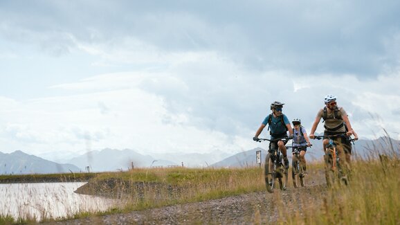 Drei Mountainbiker fahren auf einem Schotterweg am Speicherteich bei der Bürglalmbahn in Dienten | © Hochkönig Bergbahnen