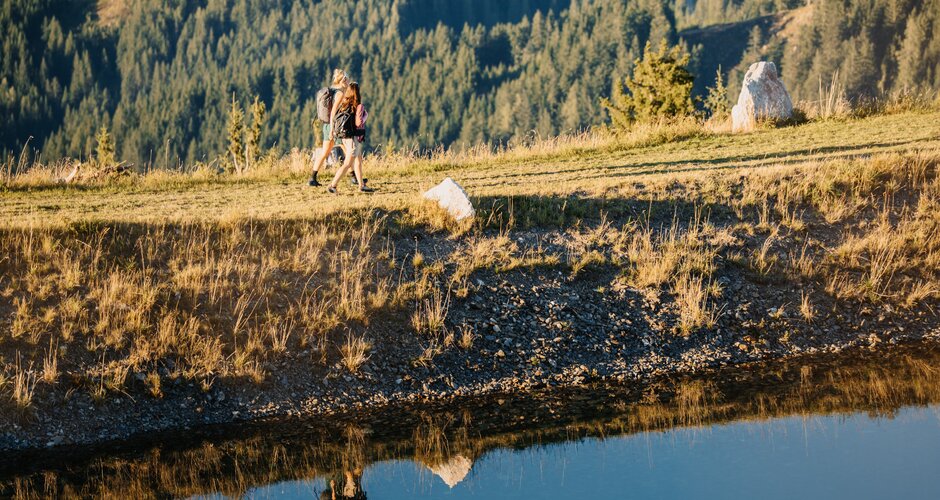Zwei Wanderer gehen am Ufer eines kleinen Teichs bei der Bürglalmbahn in Dienten über eine Wiese. | © Hochkönig Bergbahnen