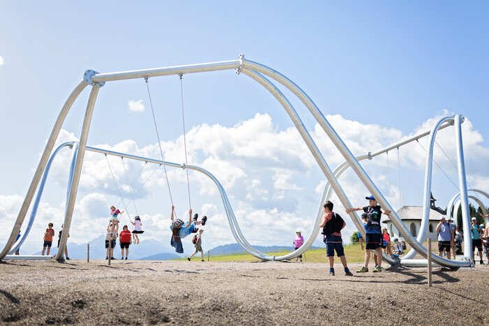 Kids swinging on oversized swings at Hochkönig swing park with Alpine backdrop on a sunny day | © Manu Lochner
