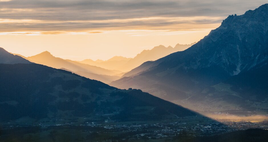 Blick über Dienten auf ein Tal mit Bergsilhouetten, Sonnenstrahlen und dicht bewölktem Himmel im Morgenlicht. | © Hochkönig Bergbahnen