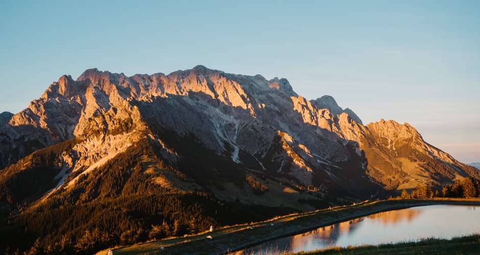 Sonnenlicht beleuchtet ein felsiges Bergmassiv über einem Speicherteich in Dienten unter klarem Himmel. | © Hochkönig Bergbahnen