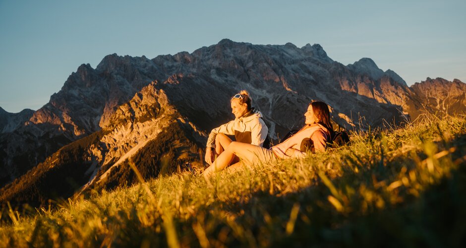 Zwei Wanderinnen sitzen im Abendlicht auf einer Wiese in Dienten vor einem felsigen Bergmassiv nahe der Gabühelbahn. | © Hochkönig Bergbahnen