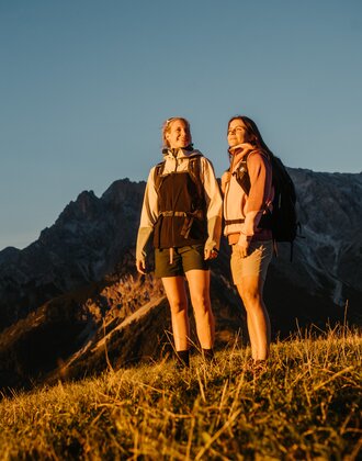 Zwei Wanderinnen stehen im Abendlicht auf einer Wiese in Dienten vor einem felsigen Bergmassiv nahe der Gabühelbahn. | © Hochkönig Bergbahnen