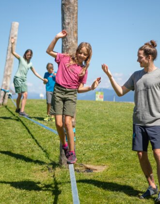 Ein Mädchen balanciert auf einer Slackline in Mühlbach, während ein Mann hilft und zwei Kinder folgen. | © Hochkönig Bergbahnen, Roland Haschka