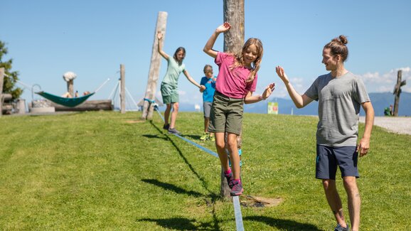 Ein Mädchen balanciert auf einer Slackline in Mühlbach, während ein Mann hilft und zwei Kinder folgen. | © Hochkönig Bergbahnen, Roland Haschka
