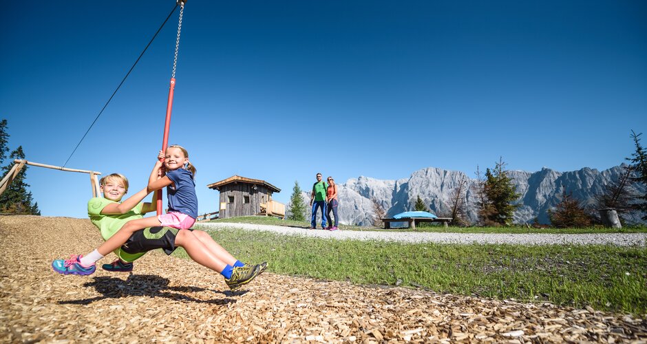 Zwei Kinder sausen lachend an der Seilbahn des Almspielplatzes entlang, im Hintergrund das Hochkönigmassiv